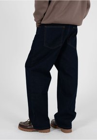 Person, die dunkle Jeans mit weitem Bein und braune Mokassin-Schuhe trägt, steht vor einem einfarbigen hellen Hintergrund.