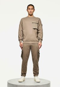 Uomo in piedi su una piattaforma rotonda che indossa una felpa taupe e pantaloni jogger abbinati con tasche a patch e sneakers bianche, sfondo neutro.