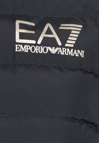 Zwarte gewatteerde vest met het reliëf zilveren logo "EA7 Emporio Armani" aan de linkerkant. Gladde textuur met horizontale stiksels.