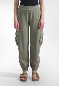 Lekkie, oliwkowozielone spodnie typu cargo z elastycznym paskiem w talii, zwężane u dołu nogawek i dwoma bocznymi kieszeniami. Noszone z brązowymi sandałami.