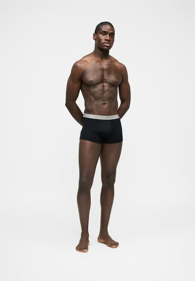 Lihaseline mees seisab paljajalu, kandes musti vormis bokserpükse halli vöökohaga, poseerides ühevärvilise hele tausta vastu.