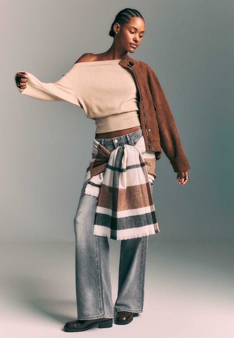 Brown suede jacket over a beige off-shoulder top, light blue jeans, and a brown striped scarf tied around the waist. Brown ankle boots.