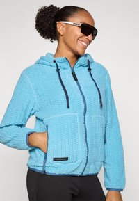 Žena s kučeravými vlasmi zaviazanými do uzla, ktorá má na sebe čierne slnečné okuliare a svetlomodrú textúrovanú mikinu na zips s rukami v vreckách, usmieva sa do boku.