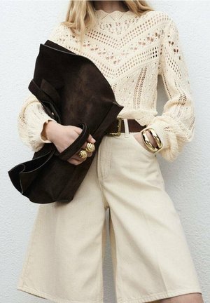 Donna che indossa un maglione lavorato a maglia color crema e pantaloni a gamba larga, con una grande pochette nera e accessoriata con anelli e braccialetto d'oro massicci.