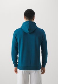 Felpa color teal con tasca a marsupio, polsini e orlo in costina. Presenta un cappuccio con cordino e un tessuto morbido e testurizzato. Mostrata vista da dietro.