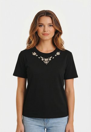 Donna che indossa una maglietta nera a maniche corte con decorazioni floreali di cristallo intorno al collo e jeans azzurri, su sfondo bianco uniforme.