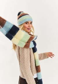 Czapka dziergana w paski niebieskie, brązowe i kremowe; wielokolorowy szalik w kratę o miękkiej fakturze; beżowy sweter z warkoczowym splotem.
