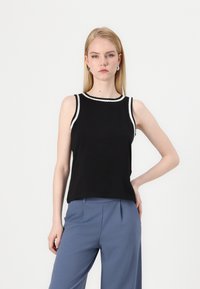 Top senza maniche nero con un bordo decorativo bianco attorno al collo, realizzato in un tessuto liscio, abbinato a pantaloni su misura azzurri.