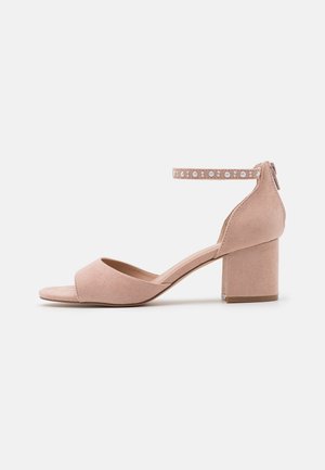 Sandales à talons en suède rose clair avec un bout ouvert, une bride de cheville ornée de clous décoratifs et un solide talon bloc. Texture lisse, design minimaliste.