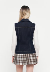 Denim prsluk u tamnoplavoj boji, tijesnog kroja i bez rukava; nosi se preko bijele košulje dugih rukava. Ispod prsluka, karirana suknja s naborima u zemljanim tonovima.