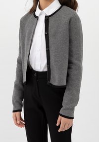 Harmaa neulottu neuletakki, jossa on pyöreä kaula-aukko, mustat reunukset ja mustat napit. Keskellä valkoista kauluksellista paitaa ja paritettu mustien housujen kanssa.