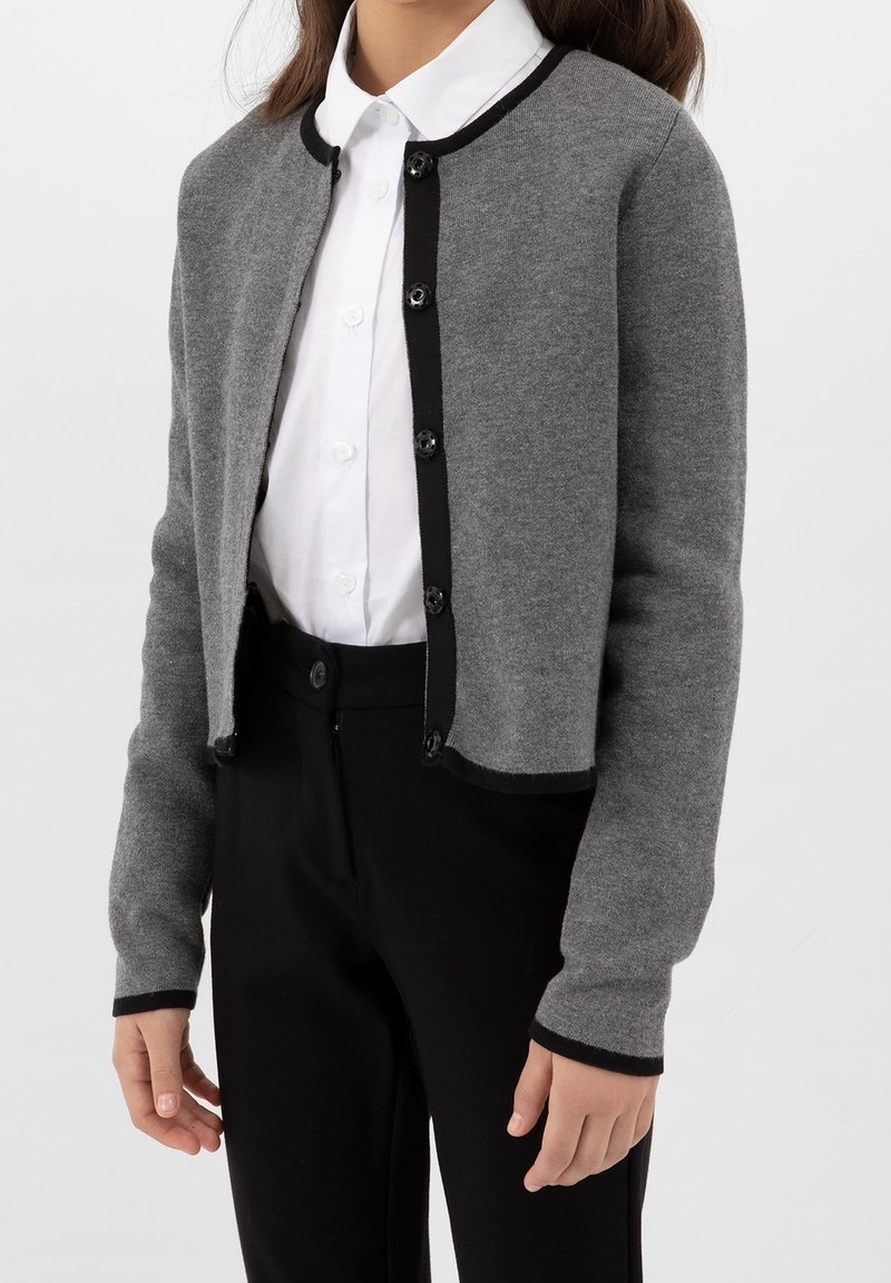 Harmaa neulottu neuletakki, jossa on pyöreä kaula-aukko, mustat reunukset ja mustat napit. Keskellä valkoista kauluksellista paitaa ja paritettu mustien housujen kanssa.