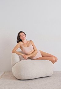 Woman in pale pink underwear reclining on a large, soft, rounded white armchair in a minimalist room with light gray carpet.