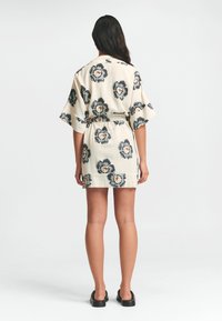 Květinový potisk krátkého kimona z béžové látky, které má široké rukávy, pás na zavazování a kontrastní tmavé květy s oranžovými středy.