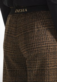 Trousers with a black and brown patterned fabric, featuring a black waistband with gold "DEHA" lettering. Textured surface with grid design.