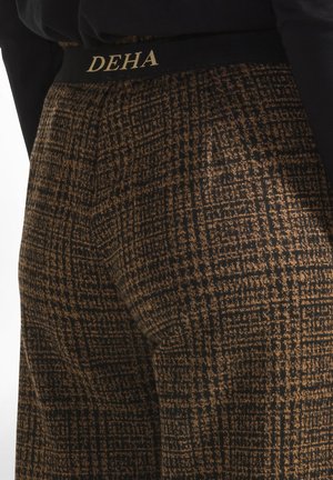 Hose aus einem schwarz-braun gemusterten Stoff, mit einem schwarzen Bund, auf dem goldene "DEHA"-Schriftzüge stehen. Strukturierte Oberfläche mit Rautenmuster.