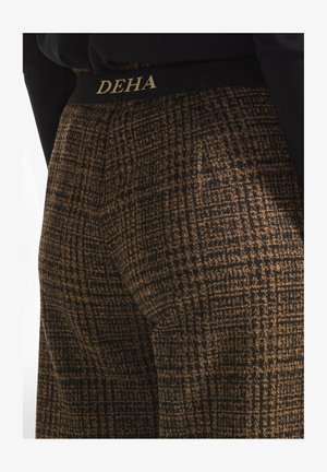 Housut, joissa on musta ja ruskea kuviollinen kangas, musta vyötärötiheys, jossa on kultaiset "DEHA"-kirjaimet. Rakenteellinen pinta, jossa on ruutukuvio.