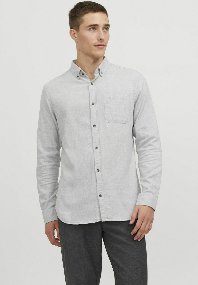 Lichtgrijze langslavige overhemd met een borstzak, voorzien van kleine bruine knopen en een button-down kraag. G gedragen met donkergrijze broeken.