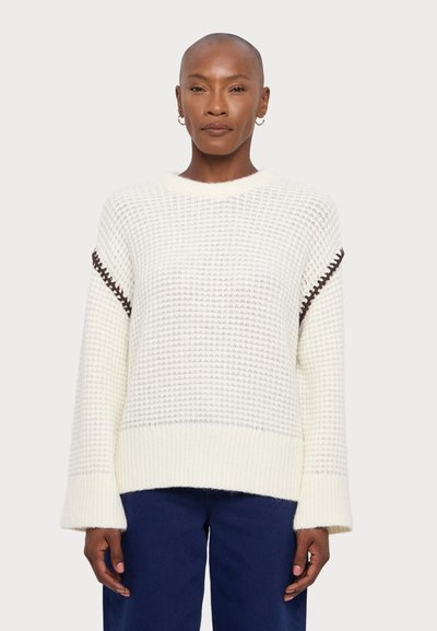Pull en tricot de couleur crème avec un motif texturé, doté de manches larges et de coutures d'accent marron foncé le long des coutures des épaules.