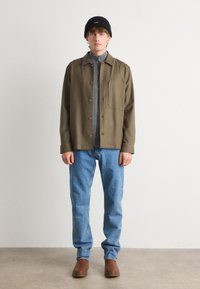 Oliven grønn skjortejakke med knapper, grå zip-up topp, blå jeans, brune støvler og en svart lue båret av en stående modell.