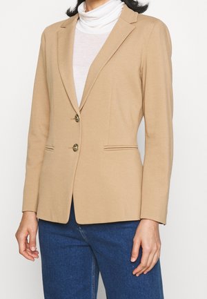 Mujer con blazer beige con dos botones dorados, cuello de tortuga blanco y vaqueros azules, mostrada desde el cuello hasta las caderas sobre un fondo liso.