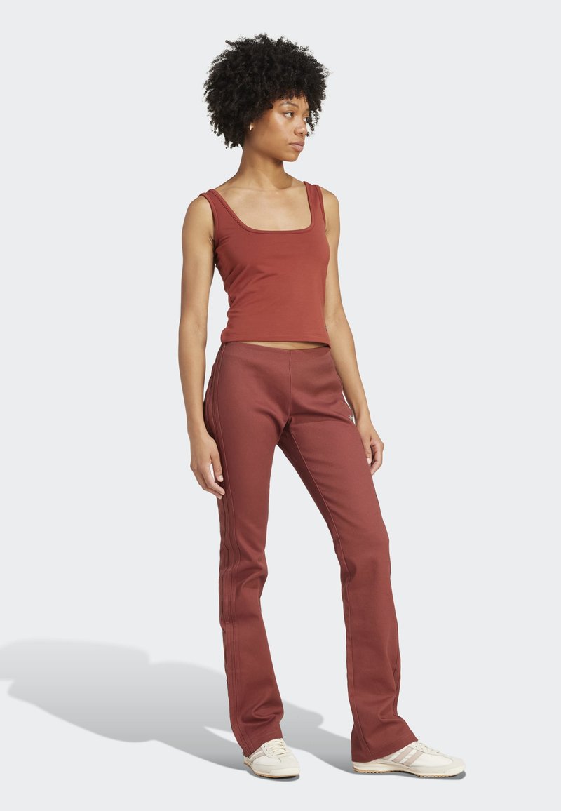 Femme aux cheveux bouclés portant un débardeur couleur rouille et un pantalon assorti, debout et regardant sur le côté sur un fond uni.