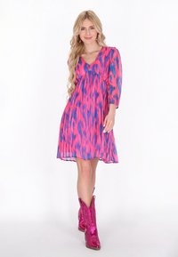 Rochie scurtă roz și albastră, cu un model, mâneci trei sferturi, decolteu în V, material fluid și talie accentuată. Pur tată cu cizme cowboy roz.
