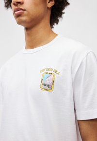 Λευκό βαμβακερό μπλουζάκι με στρογγυλή λαιμόκοψη. Διαθέτει έναν πολύχρωμο εκτυπωμένο γραφικό σχεδιασμό του Notting Hill με το κείμενο "NOTTING HILL" σε κίτρινο από πάνω.