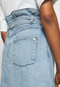 Ljusblå denimkjol med en rynkad midja, utrustad med en bakficka och silverfärgad knappdetalj. Texturen är mjuk med en n ågot sliten look.