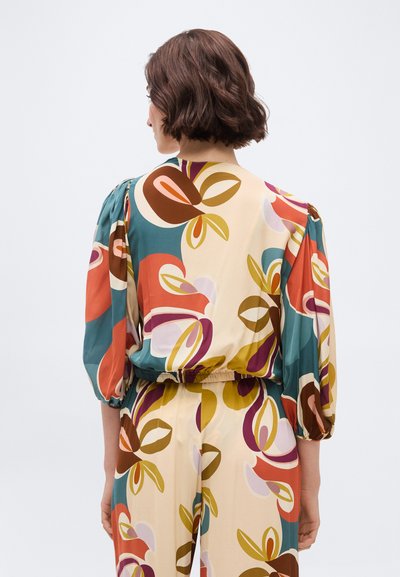 Γυναίκα με κοντά καστανά μαλλιά που φοράει πολύχρωμο μπλουζάκι με λουλουδάτο μοτίβο και αντίστοιχο παντελόνι, εμφανιζόμενη από πίσω σε απλό φόντο.