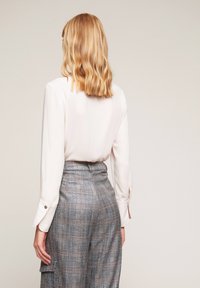 Krémová blůza s dlouhým rukávem a manžetami na knoflíky, doplněná o šedé kostkované kalhoty s vysokým pasem a boční kapsou. Hladká textura látky.