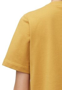Camiseta de algodón color mostaza con cuello redondo; mangas cortas y una textura suave; costuras visibles a lo largo del dobladillo y los bordes de las mangas.