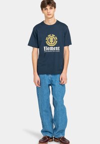 Camiseta de algodón azul marino con el logo dorado "Element" que presenta un árbol y un diseño circular, combinada con jeans de mezclilla azul claro.
