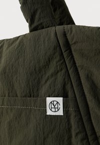 Σκούρο πράσινο υφασμάτινο υλικό με μια λευκή τετράγωνη ετικέτα ραμμένη πάνω, που απεικονίζει ένα μαύρο κυκλικό λογότυπο με επικαλυπτόμενα γράμματα.