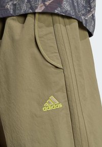 Khaki cargo shorts made from lightweight fabric, featuring yellow stitching and an embroidered Adidas logo. Includes side pockets and pleated design.