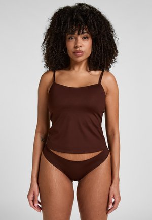 Frau mit lockigem Haar, trägt braunes Camisole und passende Unterwäsche, steht vor einfachem grauem Hintergrund, blickt nach vorne.