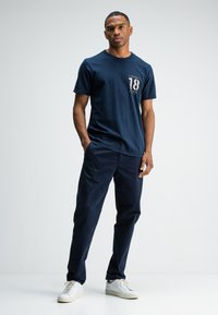 Navyblauwe katoenen t-shirt met een ronde hals en korte mouwen, met een grafisch ontwerp met het nummer 18. Gekocht met navy broeken en witte sneakers.