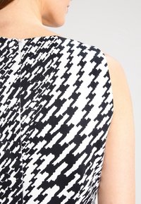 Nainen, jolla on yllään musta-valkoinen houndstooth-kuvioinen hihaton mekko, takaketjulla, näkyy takaa vaaleaa taustaa vasten.