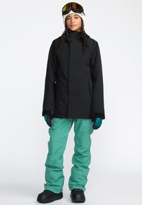 Schwarze wasserdichte Jacke, türkisfarbene Hose, schwarze Stiefel. Merkmale sind ein hoher Kragen, Taschen und Skibrille. Glatte Texturen, funktionales Design.