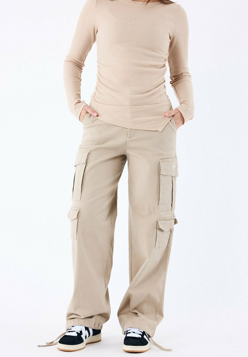 Pantalons cargo beiges avec plusieurs poches, coupe droite, poignets réglables par cordon, associés à un haut léger à côtes. Les baskets sont de couleur noire et blanche.