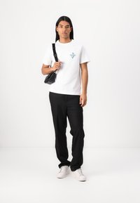 T-shirt en coton blanc avec un graphique turquoise, associé à un pantalon noir et des baskets blanches ; sac noir avec une bandoulière.