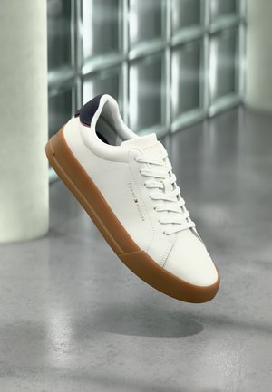 Weißer Sneaker mit niedrigem Schaft, brauner Sohle und schwarzem Fersenriemen, schwebt über einem grauen Boden in einem modernen Innenraum.