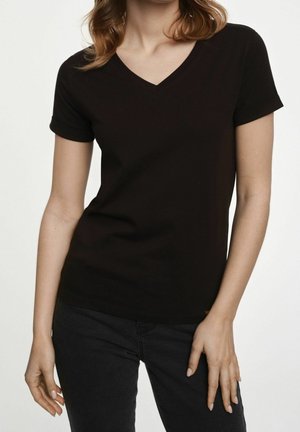 Woman wearing a fitted black V-neck short sleeve T-shirt and black pants, standing with relaxed arms against a white background.