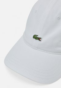 Gorra de béisbol blanca hecha de tela, presenta un logotipo de cocodrilo bordado en verde en el frente. Visera redondeada y detalles de costura visibles.