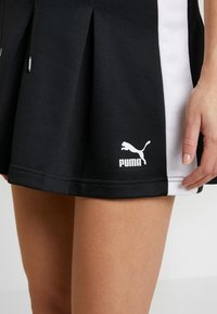 Jupe de sport plissée noir et blanc avec un tissu texturé, arborant un logo Puma blanc prominent sur l'ourlet. Coutures détaillées visibles.