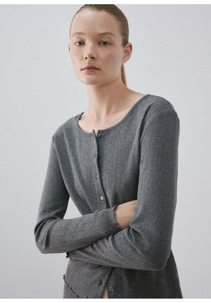 Junge Frau mit heller Haut und zurückgebundenem blondem Haar, trägt eine taillierte graue Strickjacke mit Knöpfen, steht mit verschränkten Armen.