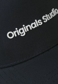 Casquette noire avec une visière cousue et un tissu texturé. "Originals Studio" est affiché en lettres blanches en relief.