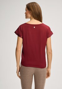 Bordeauxfarbene Kurzarmbluse mit Rundhalsausschnitt, aus weichem Stoff und einem kleinen Logo-Akzent auf dem Rücken. Schlichtes Design mit lockerer Passform.