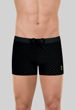 Torse masculin portant un short de bain noir à motif avec une taille à cordon et un petit logo jaune sur la cuisse gauche sur un fond uni.
