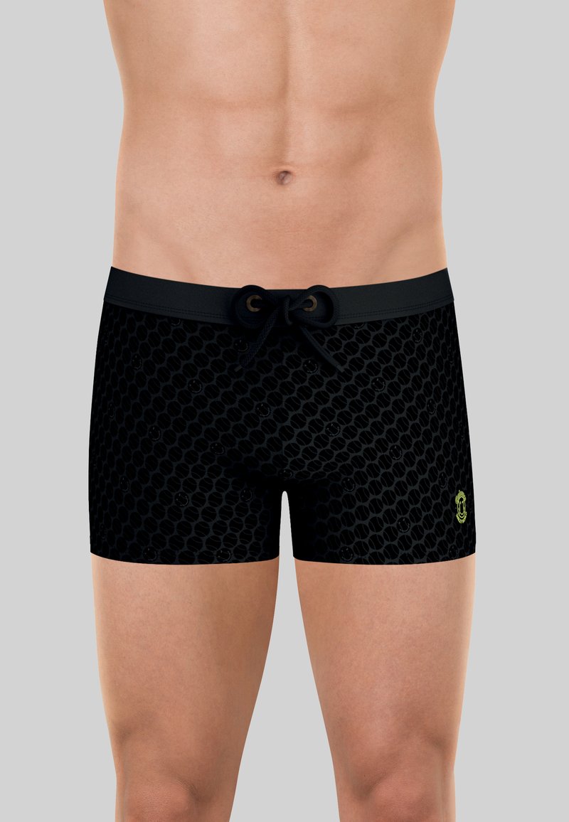 Torse masculin portant un short de bain noir à motif avec une taille à cordon et un petit logo jaune sur la cuisse gauche sur un fond uni.